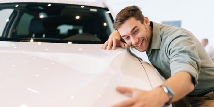 A first-time car buyer hugs the hood of his off-white SUV with a large smile on his face.